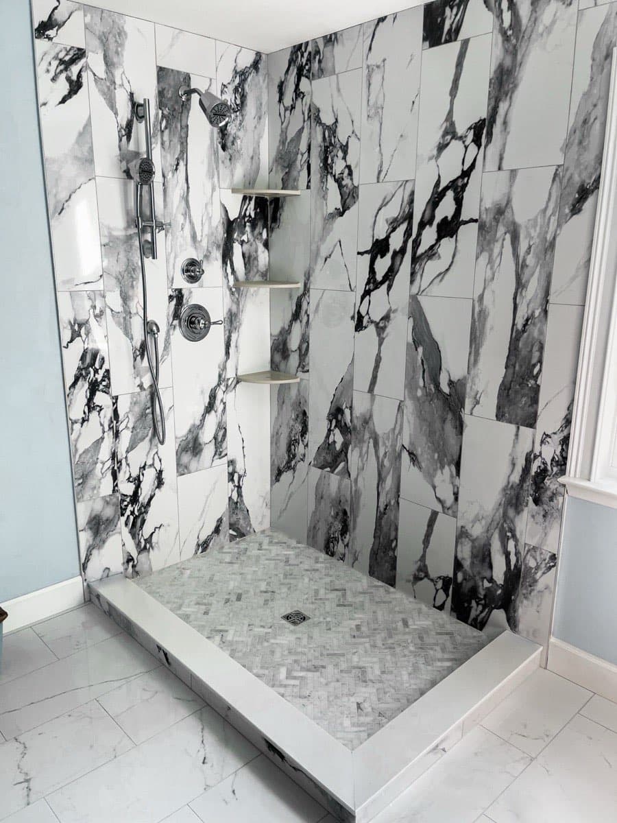 Walk-in shower with black and white marble-look wall tile, a herringbone mosaic shower pan, corner shelves, and a handheld sprayer