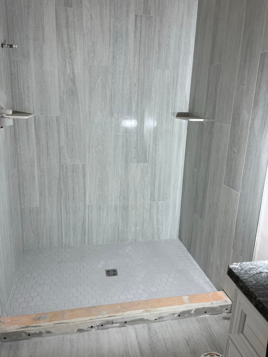 Shower with gray wood-look plank wall tile, corner shelves, and a white hexagon tile pan during installation